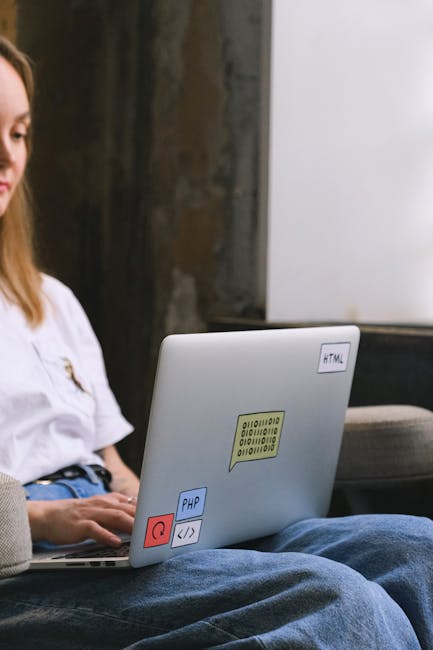 A person's hand resting on the keyboard of a silver laptop displaying lines of code or programming script on the screen. The laptop is placed on a wooden surface with a natural grain pattern. The scen